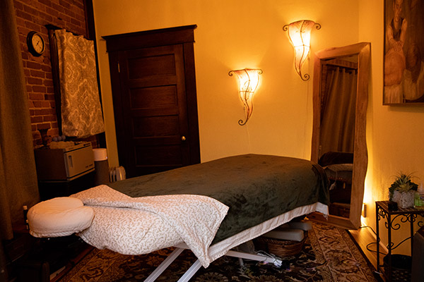 Electric massage table covered in flannel sheets and dark green blanket in treatment room with warm golden light, wood furnishings and brick wall
