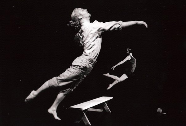 black and white photo of two dancers leaping in air with a wooden bench in background