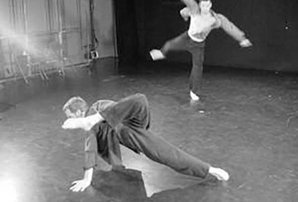 Two male dancers in studio theater, one upstage jumping, the other downstage in crouch